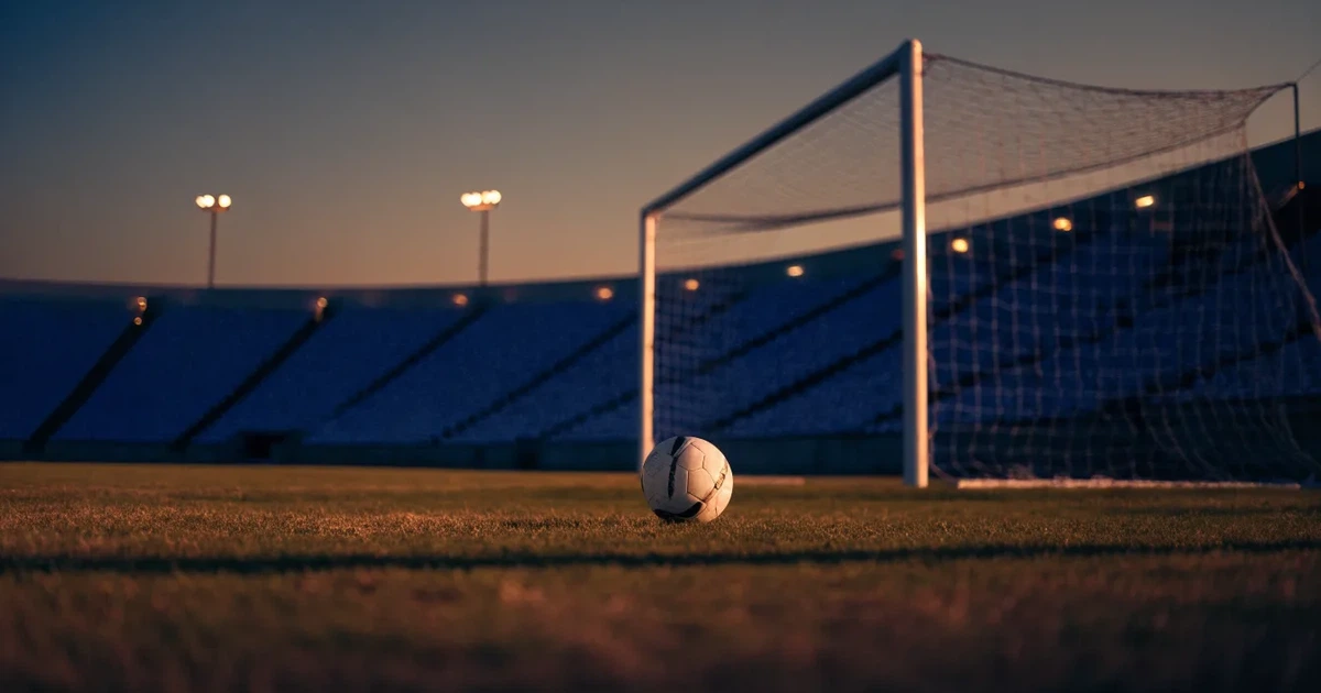 Ein Fußball liegt im Strafraum direkt vor dem Tor mit Netz, im Hintergrund leere Ränge bei Dämmerung.