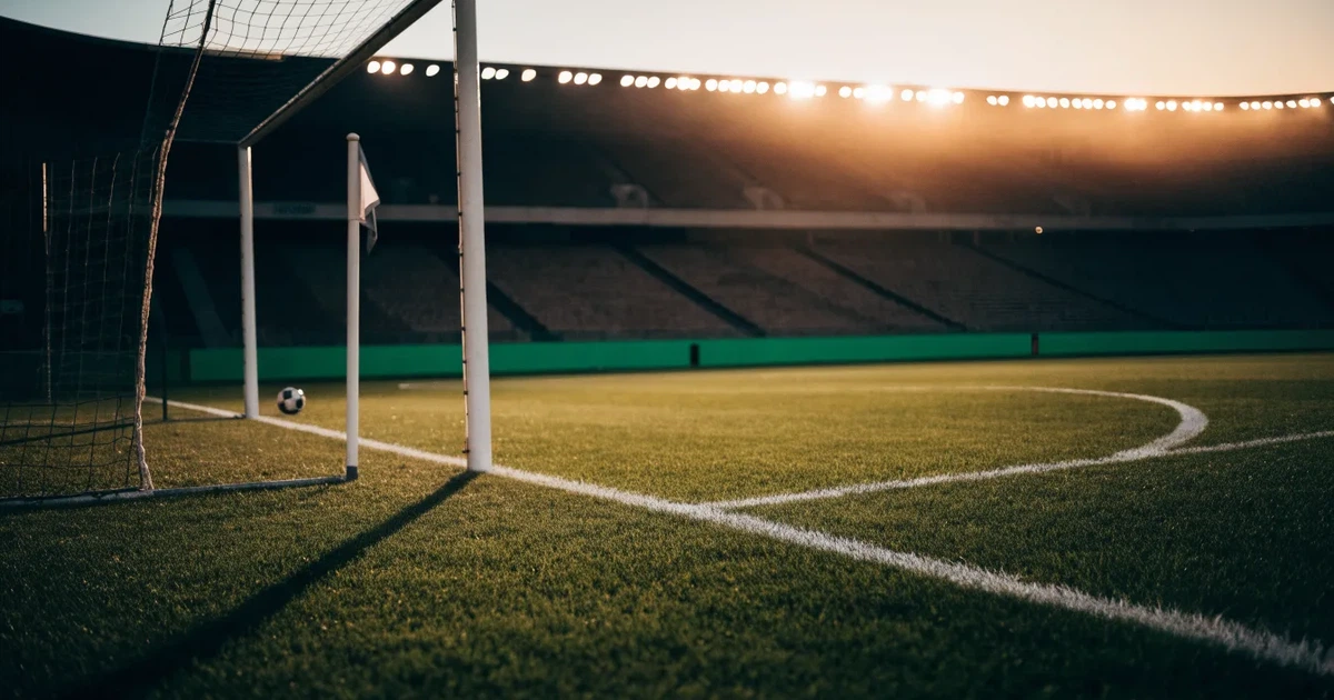 Blick durch ein Stadiontor auf einen grünen Rasen im Abendlicht, ein Ball ruht an der Eckfahne, die Tribünen sind leer.