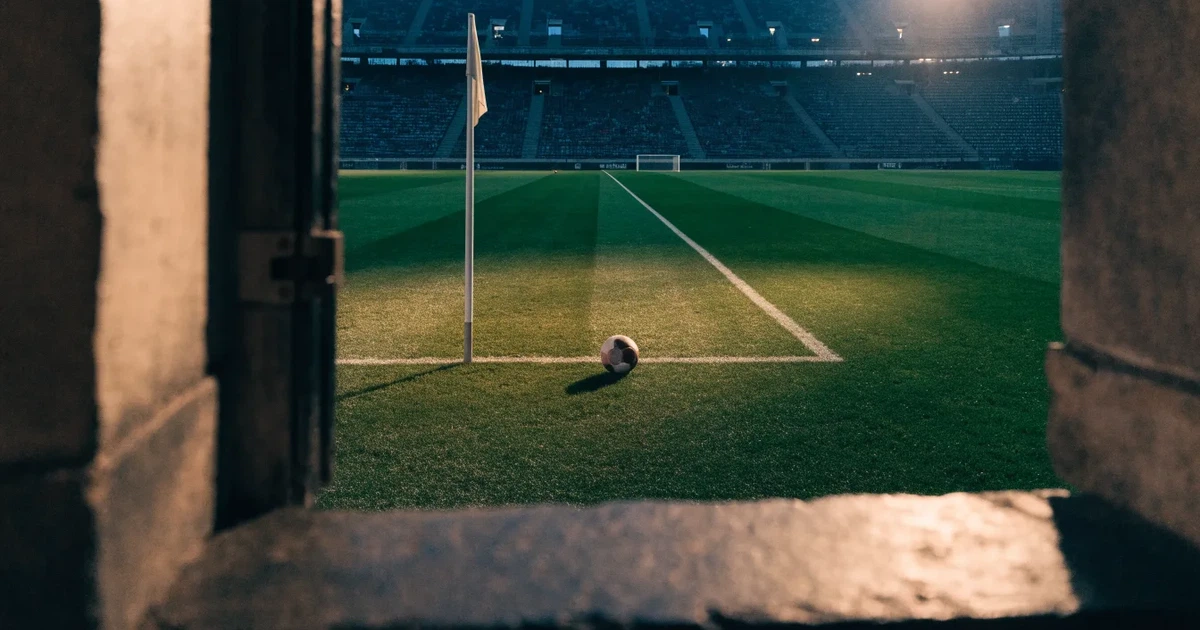 Blick durch ein Stadiontor auf einen grünen Rasen im Abendlicht, ein Ball liegt an der Eckfahne, die Ränge sind leer.
